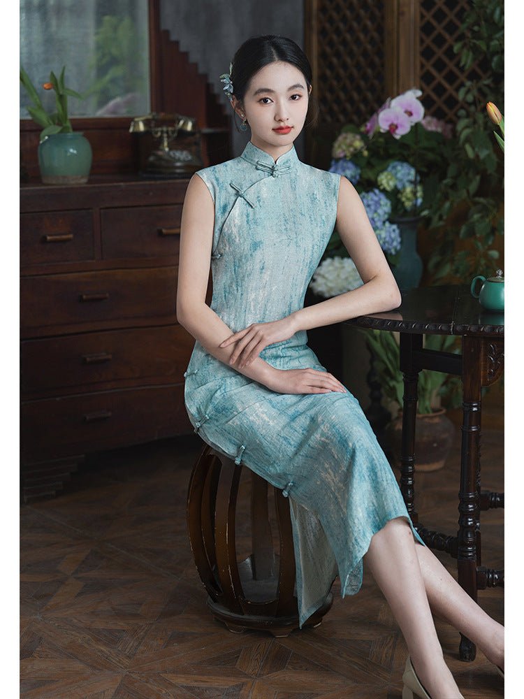 Elegant woman in a teal jacquard qipao sitting gracefully in a vintage wooden chair beside a vase of flowers.
