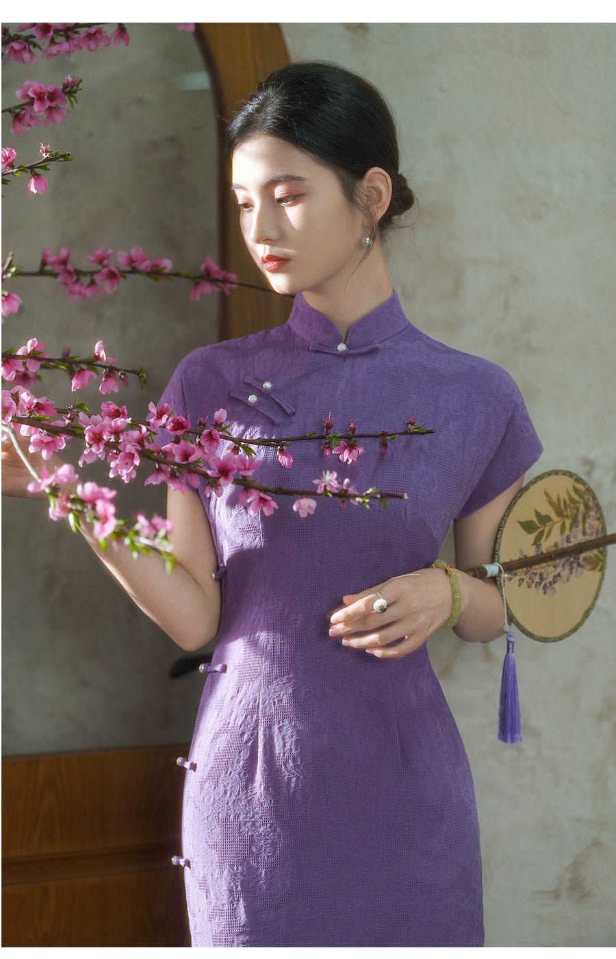 Woman wearing a classic purple qipao, holding a fan and smiling softly among peach blossoms.