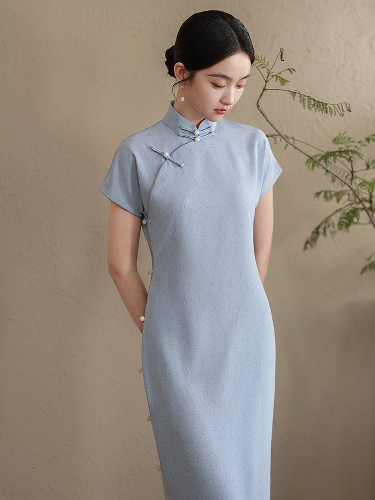 Full view of a woman standing in a pastel blue cheongsam dress with short sleeves and a calm expression.