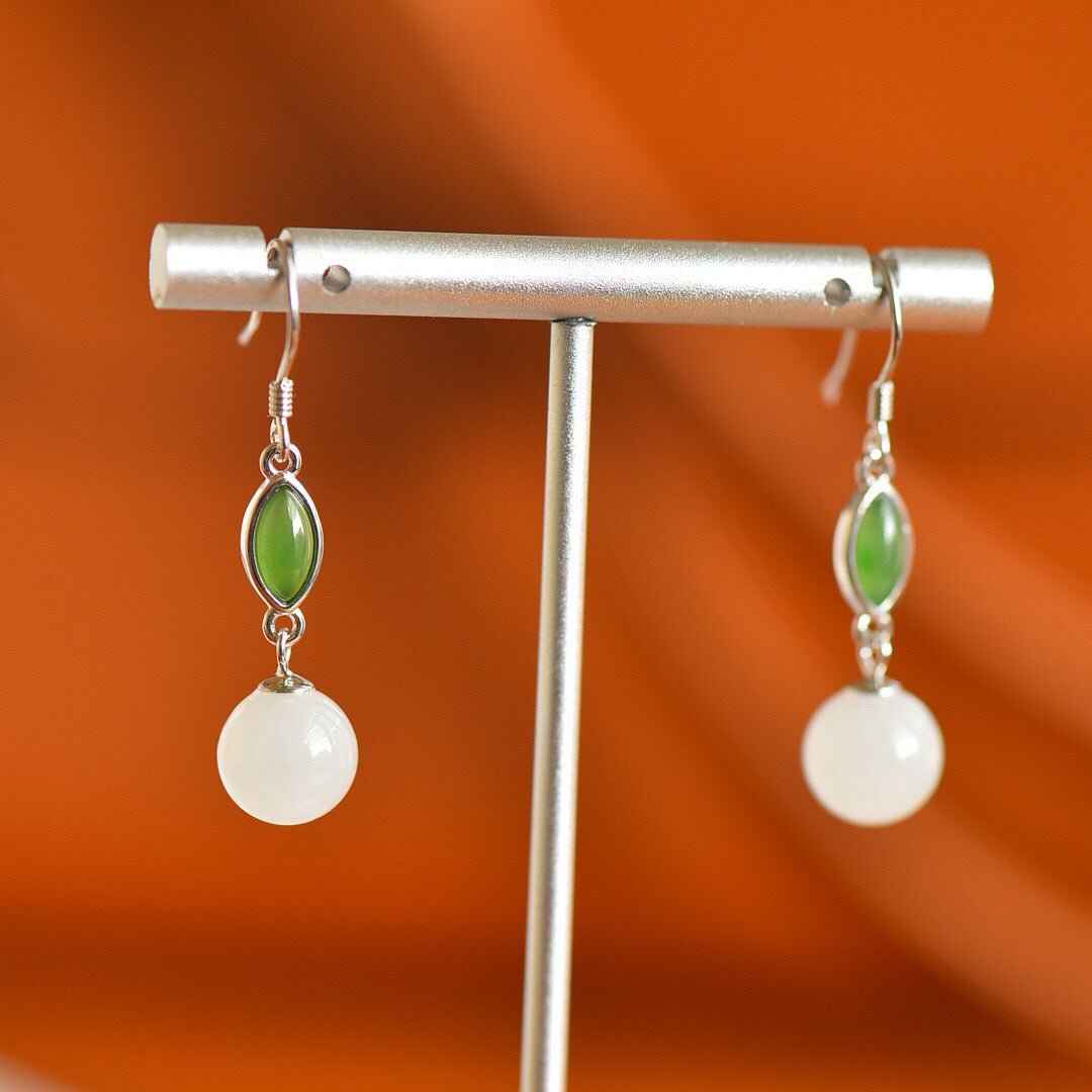 White jade and green gemstone earrings hanging from silver hooks, set against warm orange background.