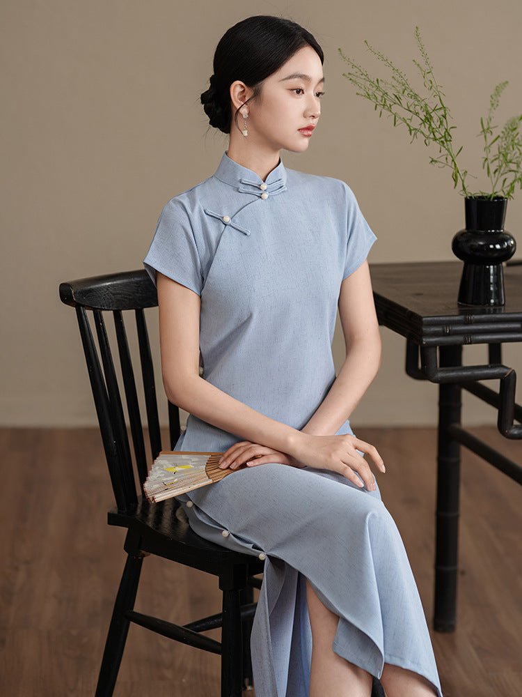 Elegant woman sitting on a wooden chair in a soft blue Chinese-style qipao with floral embroidery.