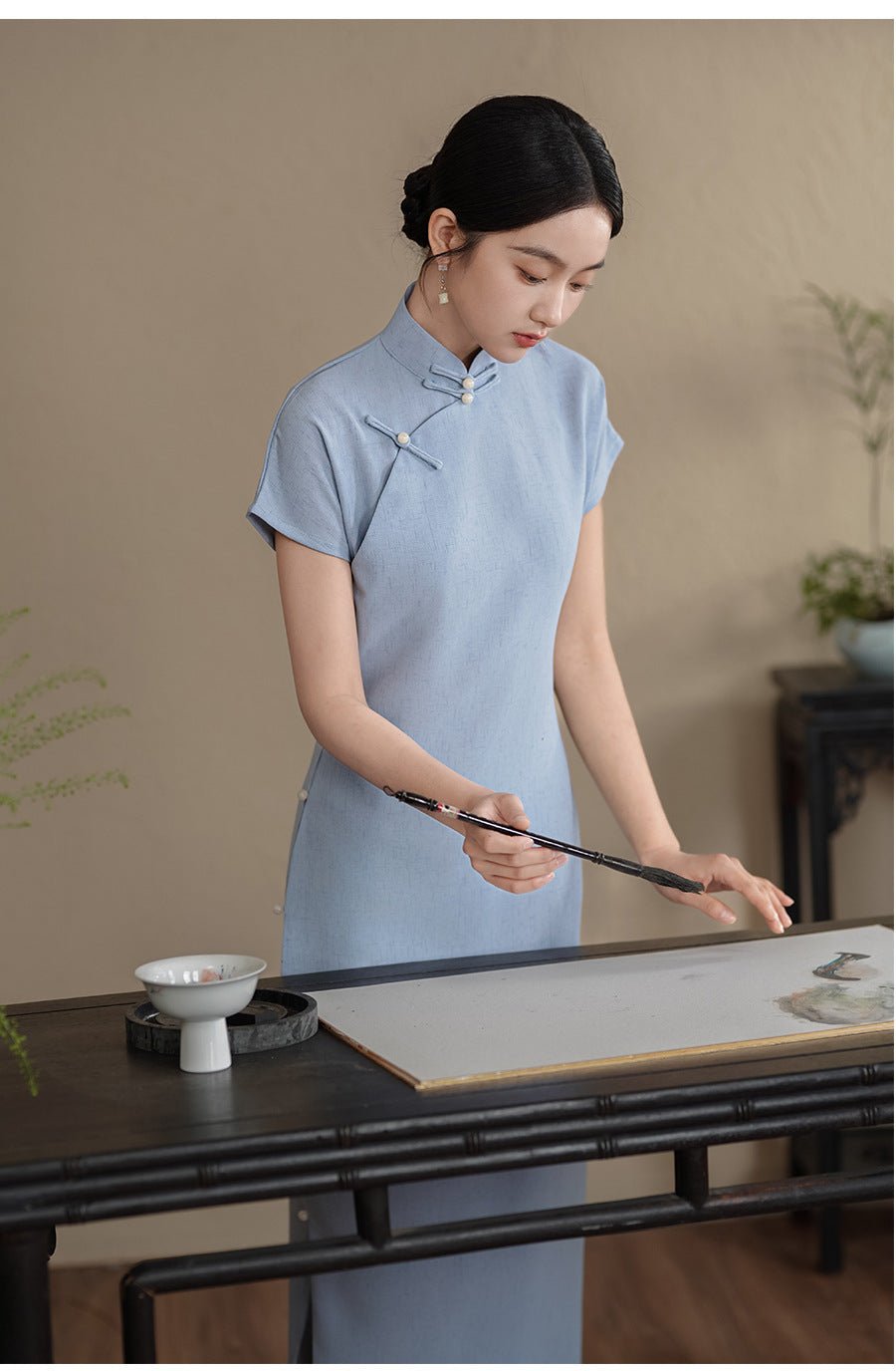 Woman wearing a light blue qipao dress with traditional stand collar, standing by a wooden table.