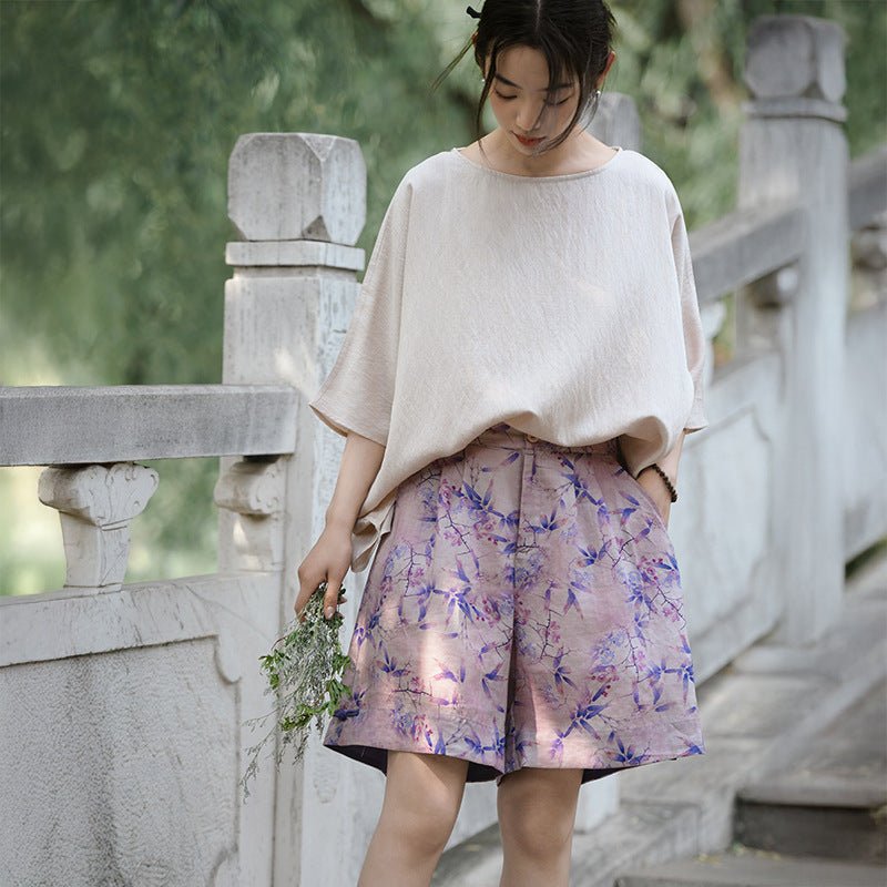 Woman in soft cream top and purple floral shorts standing on a stone bridge – peaceful summer vibe